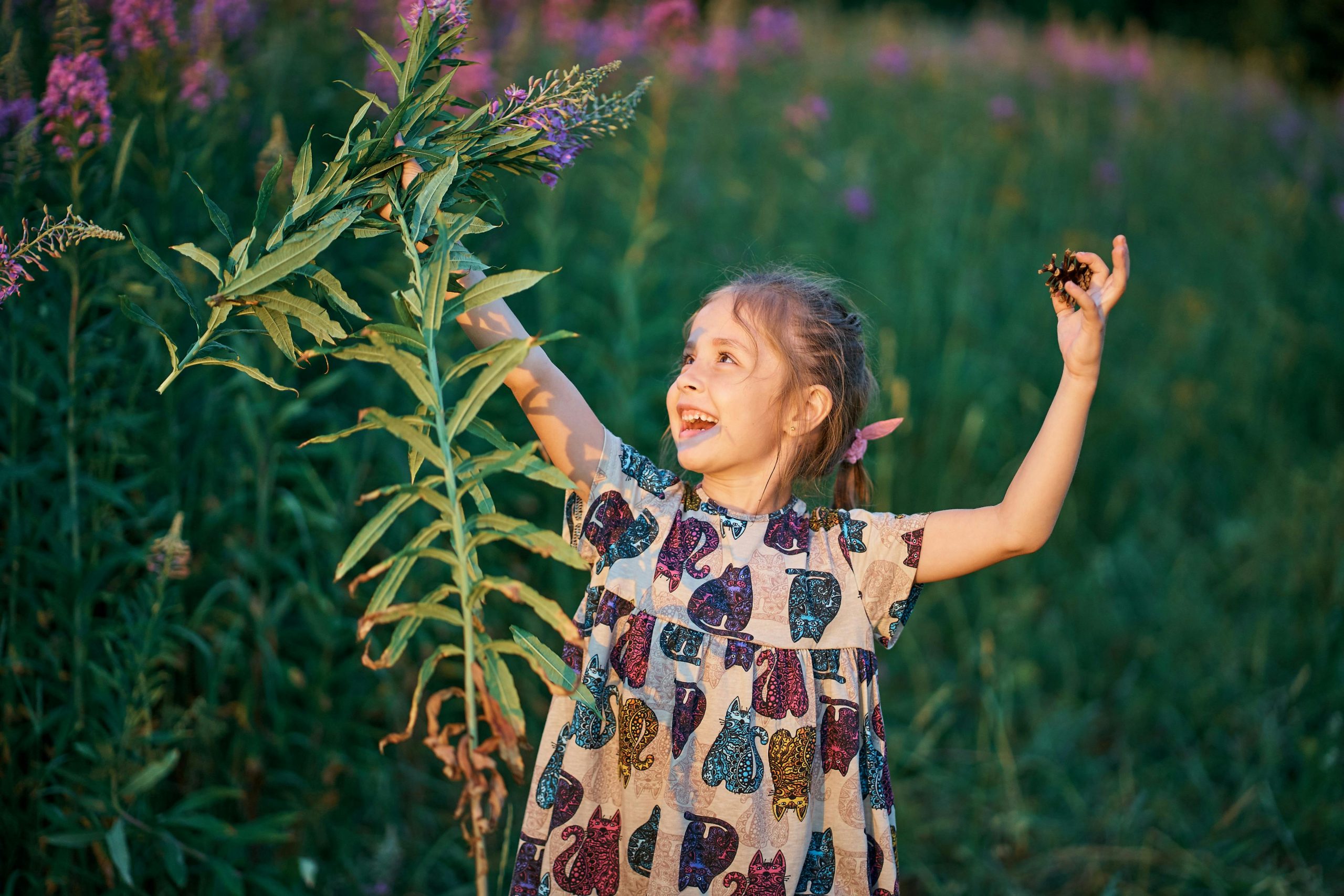 A young girl joyfully playing among tall green plants and purple flowers, enjoying a sunny day outdoors.